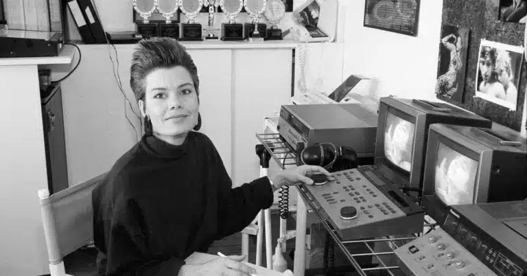 Candida Royalle, cheveux courts et grandes boucles d'oreilles, assise devant ses moniteurs de montage, trophées au mur, photo en noir et blanc
